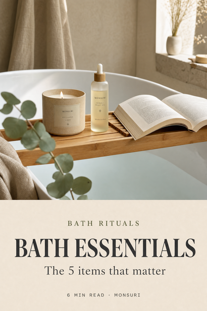 Bath essentials cover — bamboo bath tray bridging a tub holding a soy candle, body oil, and an open book; magazine cover composition for Monsuri Journal