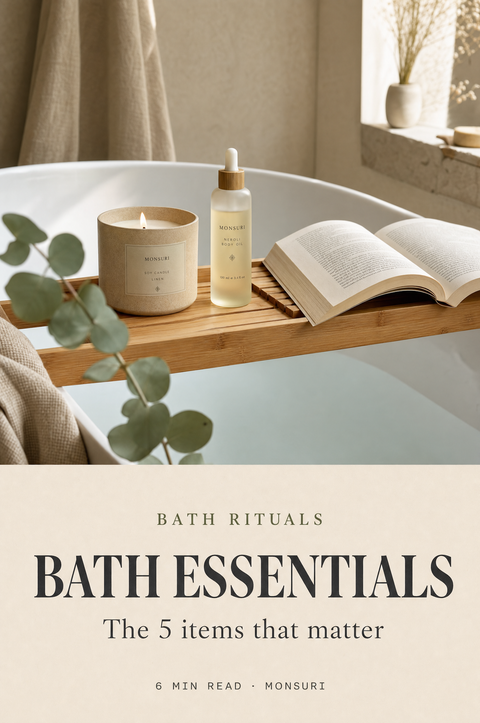 Bath essentials cover — bamboo bath tray bridging a tub holding a soy candle, body oil, and an open book; magazine cover composition for Monsuri Journal