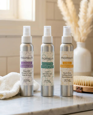 The three Monsuri shower sprays standing on a marble bathroom counter beside a folded towel and a dry brush, showing the size of the 4 oz bottles in a real bathroom