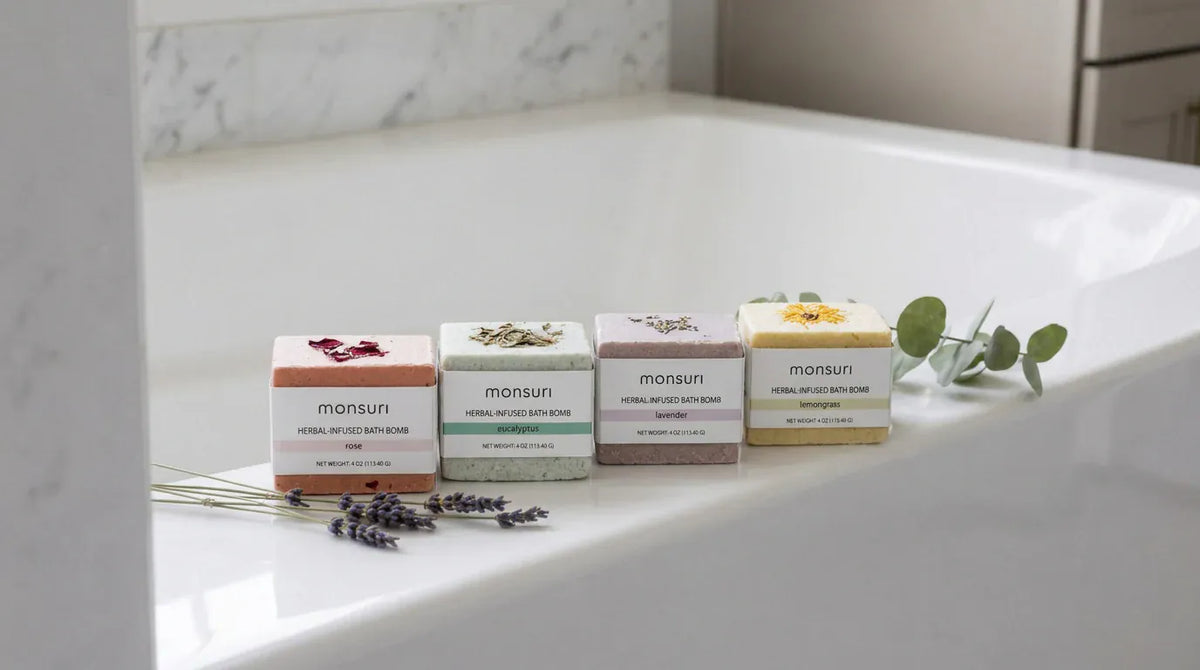 The four bath bombs standing in a row on the rim of a white tub in bright daylight, with dried lavender stems and a eucalyptus sprig laid beside them.