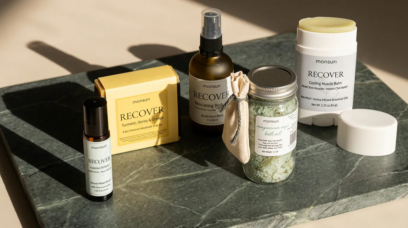 The five-piece muscle relief set arranged across a green marble slab in low sun — the nourishing body oil, the magnesium sage bath salts with their muslin bag, the cooling muscle balm stick with its cap off beside it, the boxed turmeric, honey and orange soap and the essential-oil roller.