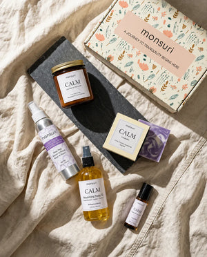 All five pieces of the Calm Moments Sanctuary set laid on cream linen with the botanical kraft gift box - the CALM lavender-chamomile candle on slate, the lavender relaxing shower spray, the lavender soap bar, the CALM nourishing body oil and the pocket essential oil roller
