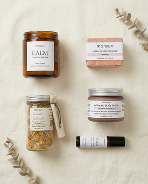 Flat lay of the five-piece lavender bath gift set on cream linen: the CALM lavender chamomile candle, the herbal-infused lavender bath bomb, the lavender bath salt jar, the lavender whipped body butter and the CALM essential oil roller, with dried eucalyptus alongside.