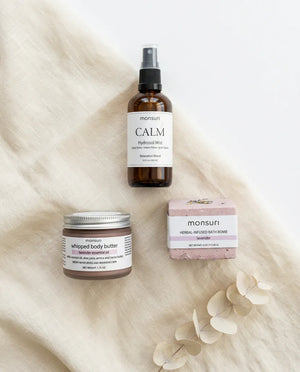 Flat lay of the Evening Wind Down bedtime trio on cream linen: the Calm Lavender Hydrosol Mist spray bottle, the lavender whipped body butter jar and the tranquil lavender bath bomb, with dried eucalyptus alongside.