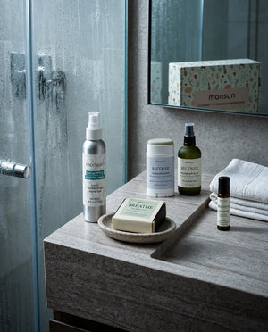 The eucalyptus shower spray, balm stick, body oil, soap and roller on a bathroom counter beside a steamy shower door