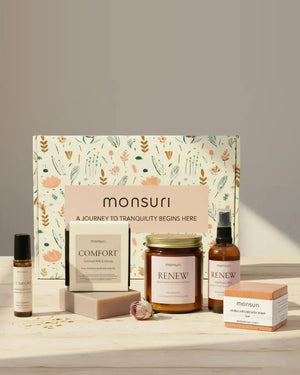 All five pieces of the Monsuri Hug in a Box sympathy gift with the botanical gift box - the 7 oz RENEW rose sandalwood candle, the COMFORT oatmeal milk and honey soap bar, the rose geranium hydrosol mist, the herbal-infused rose bath bomb and the COMFORT essential oil roller, on pale wood with rose petals