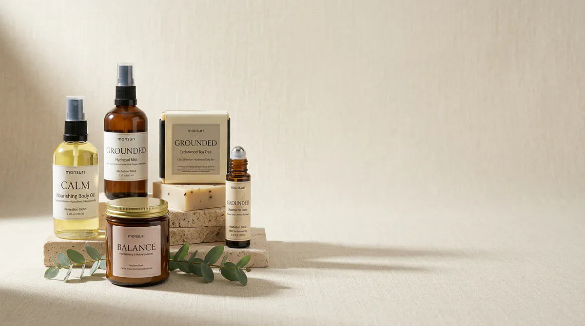 All five mindfulness pieces grouped on travertine blocks with eucalyptus at their base — the CALM body oil, the GROUNDED hydrosol, the boxed cedarwood tea tree soap, the BALANCE palo santo candle and the meditation roller — against a pale plaster wall.