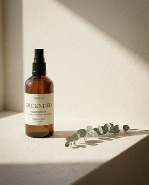 The GROUNDED hydrosol mist alone in its amber glass spray bottle on a pale plaster ledge, a eucalyptus stem laid flat beside it in hard afternoon light.