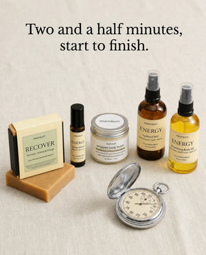 Two and a half minutes, start to finish: the whole five-piece energizing routine laid out on linen beside a stopwatch — the RECOVER turmeric soap, the energy roller, the lemongrass-rosemary body butter, the citrus hydrosol mist and the nourishing body oil.