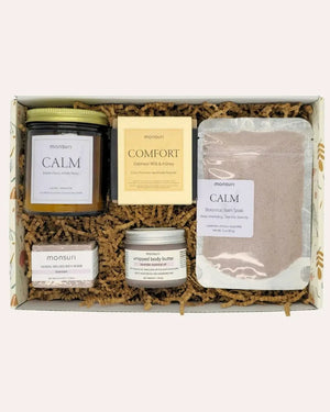 Mom's Wind-Down Ritual arranged inside its botanical kraft gift box on kraft shred, seen from above: the CALM lavender chamomile candle, the COMFORT oatmeal milk and honey soap bar, the CALM botanical bath soak pouch, the herbal-infused lavender bath bomb and the lavender whipped body butter.