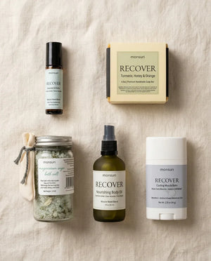 Overhead flat lay of the five-piece muscle recovery gift set on cream linen — the essential-oil roller and the boxed turmeric, honey and orange soap above; the magnesium sage bath salts with their muslin bag, the nourishing body oil in olive glass and the cooling muscle balm stick below.