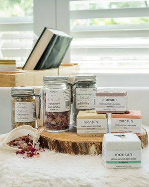 The Serenity Now bath bomb and salts combo on a wooden slice: the lavender and rose herbal bath salt jars with a muslin steeping bag of rose salts, alongside the herbal-infused lemongrass bath bomb.