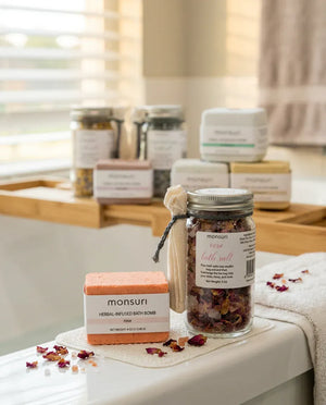 The rose bath bomb and the rose herbal bath salt jar resting on the rim of a tub beside a folded towel, with the rest of the set on a wooden bath caddy behind and scattered rose petals.