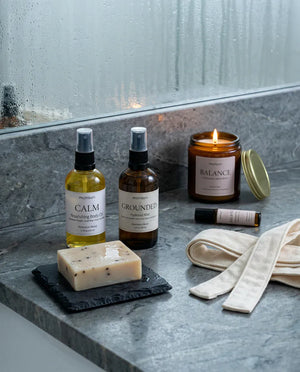 The set arranged on a stone bath ledge at dusk - the lit palo santo candle, the body oil and hydrosol mist, the soap on slate and the roller beside a folded towel