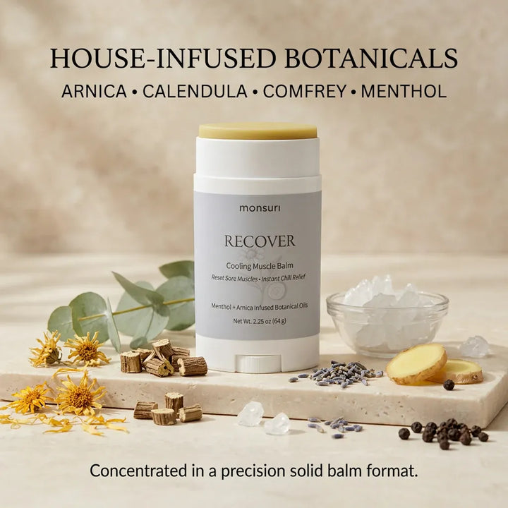 Monsuri RECOVER cooling muscle balm stick surrounded by its house-infused botanicals -- arnica flowers, wood chips, lavender, ginger root, and menthol crystals -- on a stone slab.