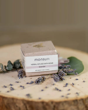 Close-up of the Monsuri lavender herbal-infused bath bomb, 4 oz, on a wood slice scattered with dried lavender buds and eucalyptus