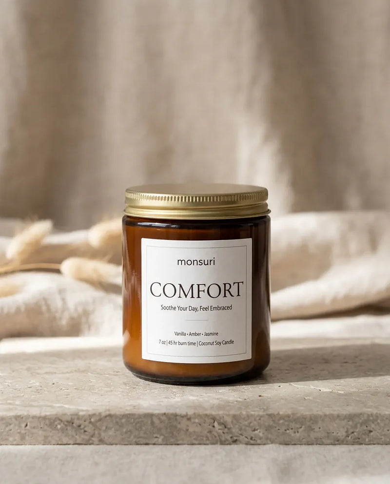 Monsuri COMFORT vanilla amber jasmine candle with its brass lid on, styled on a stone ledge against linen drapery and dried wheat stalks.