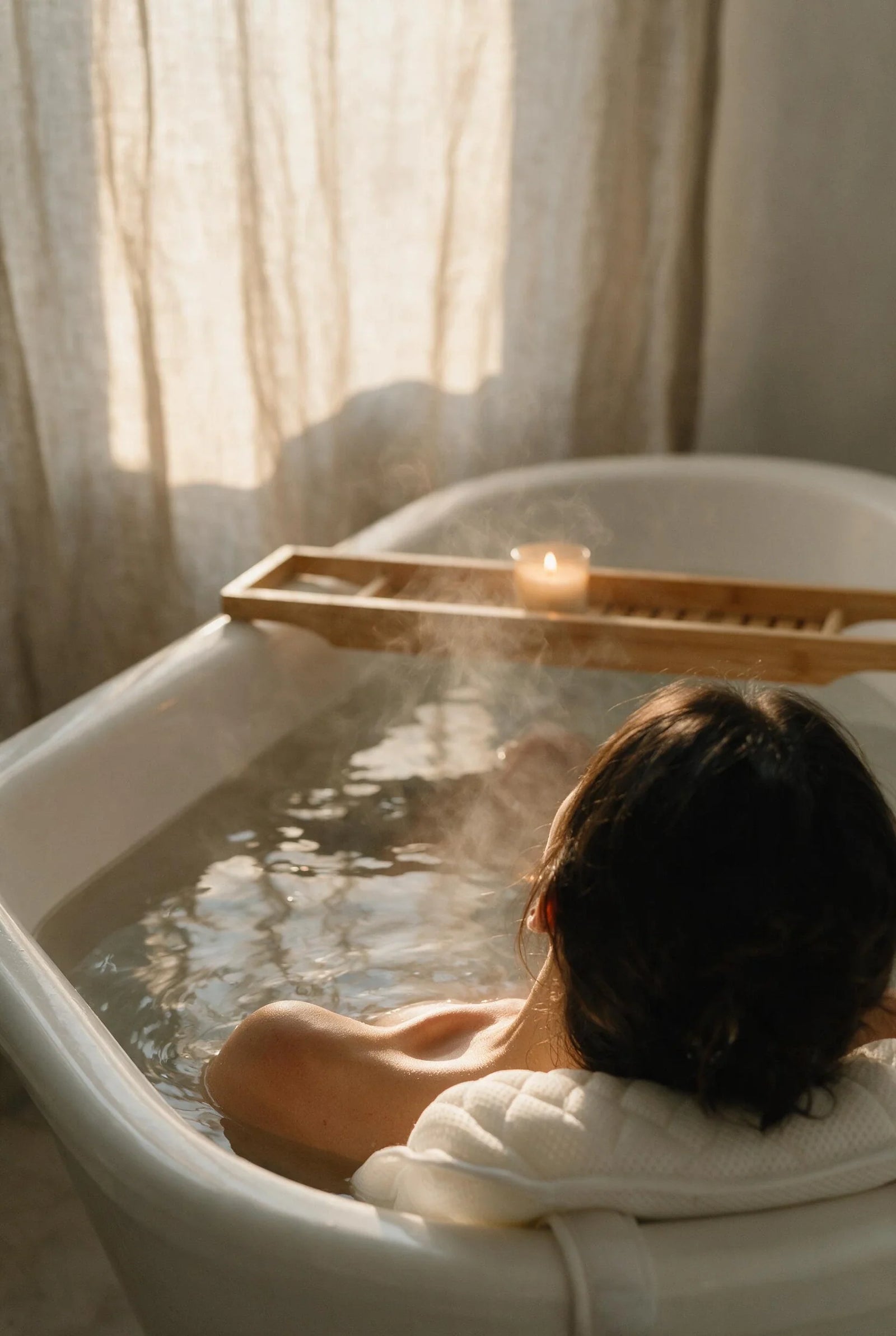 Bath pillow for head and neck in use — shot from behind a filled tub, a woman reclines with her head and neck resting on a white quilted bath pillow, steam rising past a bamboo tray and a lit candle.