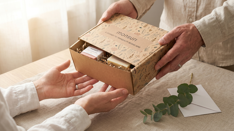 Husband-gifting moment — handing the Monsuri Romantic Spa Gift Set kraft gift box to his wife on her birthday, hands-only intimate framing, the thoughtful birthday gift for wife hero.