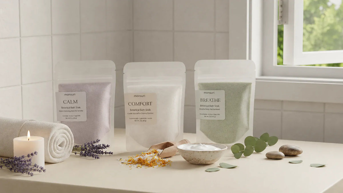 The three Monsuri bath soaks along a bathroom counter — CALM in lavender, COMFORT in coconut milk and BREATHE in eucalyptus, each 3 oz — with a lit candle, a rolled towel, dried lavender, calendula petals and eucalyptus leaves.