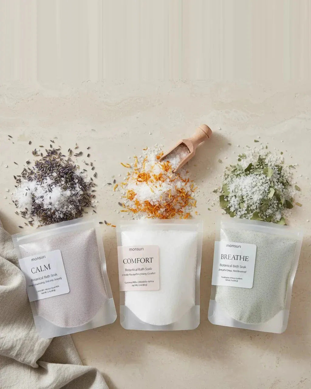 The three bath soaks with their botanicals laid out beside them: dried lavender for CALM, calendula petals and a wooden scoop for COMFORT, and eucalyptus for BREATHE.