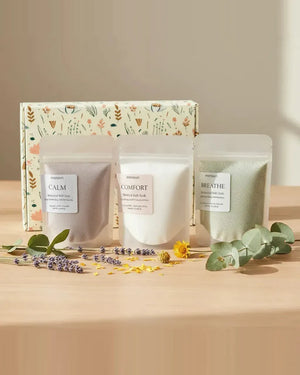 The three 3 oz bath soaks — CALM lavender, COMFORT coconut milk and BREATHE eucalyptus — on a wooden table with dried lavender, calendula petals and eucalyptus sprigs, a botanical-print gift box standing behind them.
