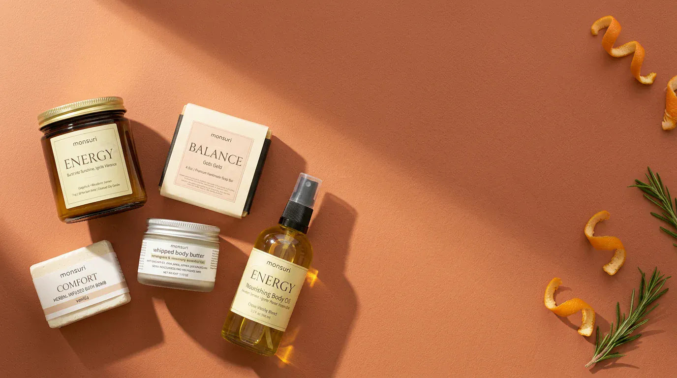 All five pieces grouped at one end of a terracotta surface with orange peel and rosemary trailing away from them — the ENERGY candle, the boxed BALANCE Gobi Gold soap, the COMFORT bath bomb, the whipped body butter and the ENERGY body oil.