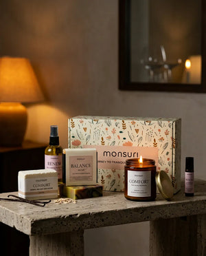 The Birthday Bliss set on a stone console in a warm bedroom at night, the COMFORT candle lit beside the body oil, soap, bath bomb and roller