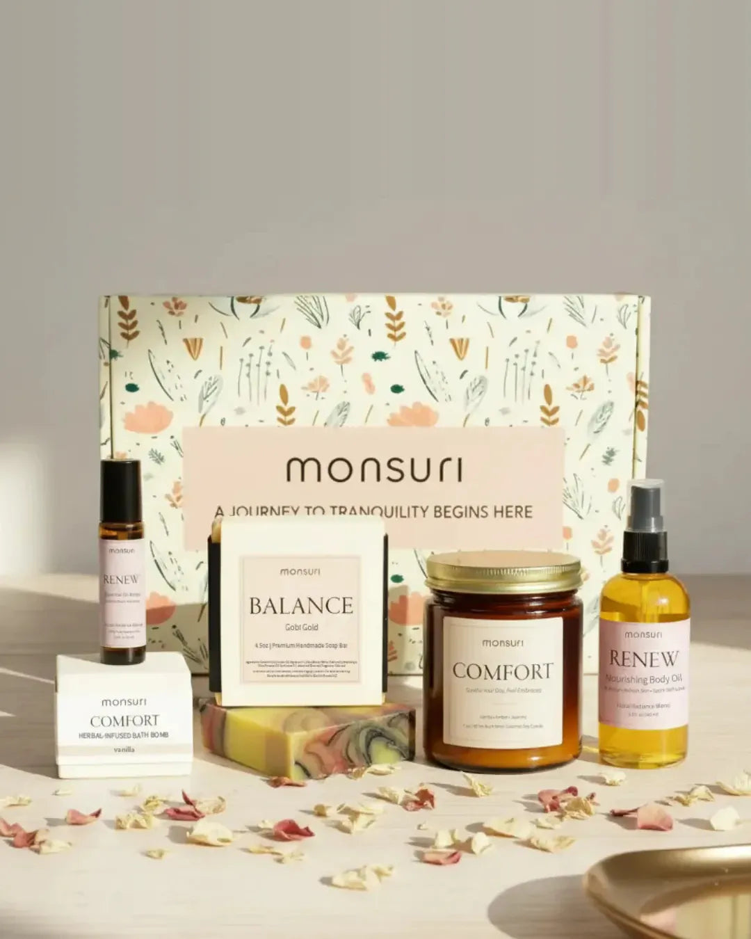 All five pieces of the Birthday Bliss celebration set with the Monsuri botanical gift box - the RENEW Floral Radiance roller, the COMFORT vanilla bath bomb, the BALANCE Gobi Gold soap bar, the 7 oz COMFORT vanilla amber jasmine candle and the RENEW Floral Radiance body oil, scattered with rose petals