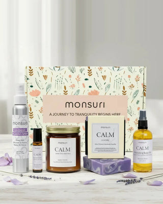 The Calm Moments Sanctuary five-piece lavender gift set with the monsuri botanical box - the 4 oz lavender relaxing shower spray, the essential oil roller, the CALM lavender-chamomile candle, the 4.5 oz lavender soap bar and the CALM nourishing body oil, with lavender sprigs and petals on a white table