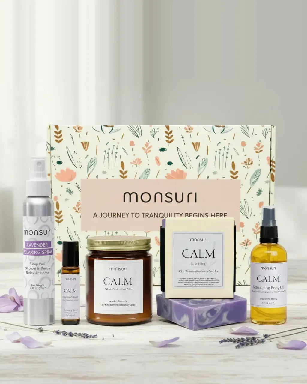 The Calm Moments Sanctuary five-piece lavender gift set with the monsuri botanical box - the 4 oz lavender relaxing shower spray, the essential oil roller, the CALM lavender-chamomile candle, the 4.5 oz lavender soap bar and the CALM nourishing body oil, with lavender sprigs and petals on a white table
