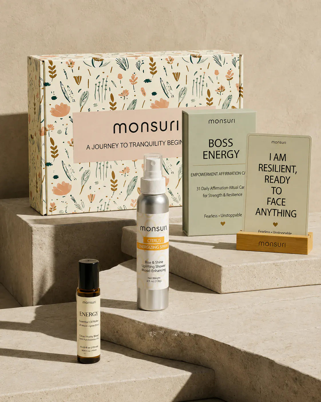 Confidence and Empowerment gift set on travertine steps in front of the Monsuri botanical gift box - BOSS ENERGY deck of 31 empowerment affirmation-ritual cards, a card reading I AM RESILIENT READY TO FACE ANYTHING in its wooden stand, the 4 fl oz citrus energizing shower spray and the ENERGY essential oil roller