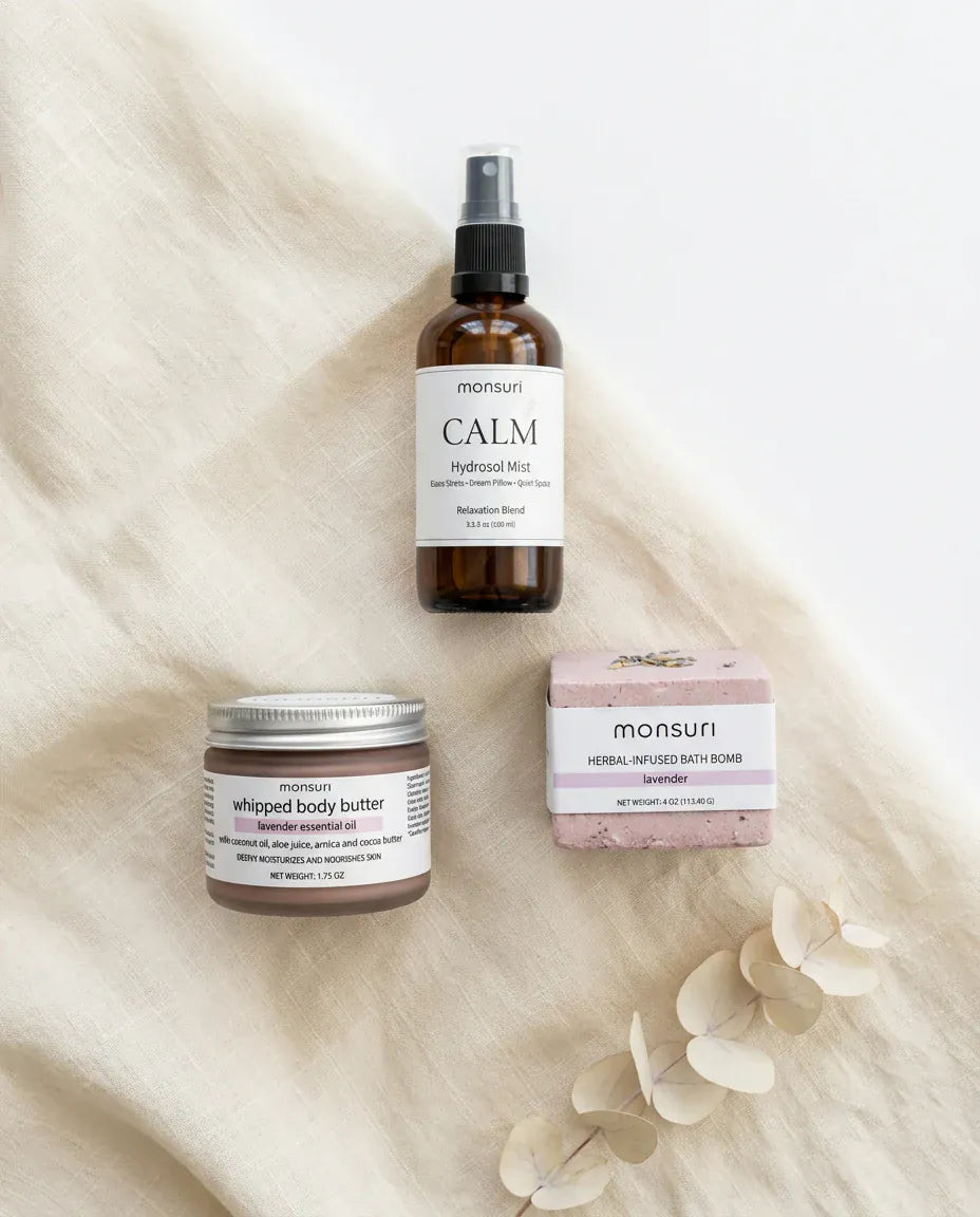 Flat lay of the Evening Wind Down bedtime trio on cream linen: the Calm Lavender Hydrosol Mist spray bottle, the lavender whipped body butter jar and the tranquil lavender bath bomb, with dried eucalyptus alongside.