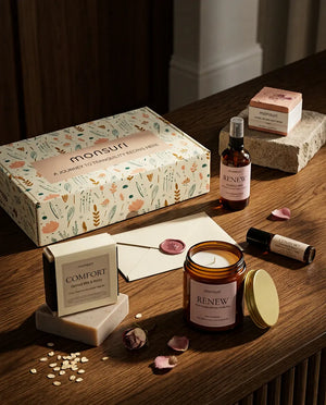 The gift box open on a wooden table beside a card and envelope, the lit candle, the soap and the roller, in the moment of writing the note