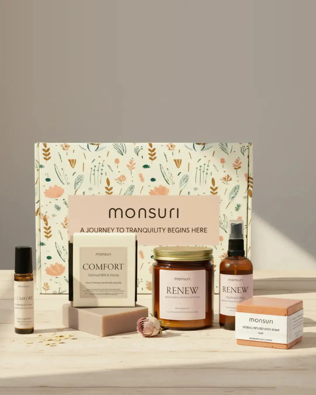 All five pieces of the Monsuri Hug in a Box sympathy gift with the botanical gift box - the 7 oz RENEW rose sandalwood candle, the COMFORT oatmeal milk and honey soap bar, the rose geranium hydrosol mist, the herbal-infused rose bath bomb and the COMFORT essential oil roller, on pale wood with rose petals