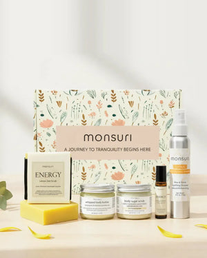 The five pieces of the Little Box of Sunshine gift in front of the Monsuri botanical box - the ENERGY lemon zest soap bar, whipped body butter, body sugar scrub, ENERGY essential oil roller and the CITRUS energizing shower spray, with yellow petals on pale wood