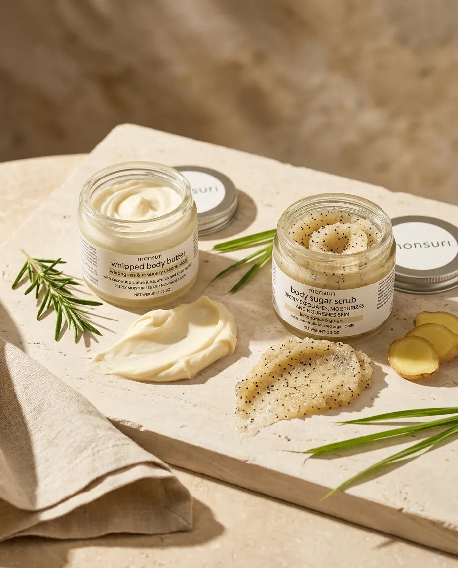 The lemongrass rosemary whipped body butter and the lemongrass ginger body sugar scrub open side by side on stone, a swirl of each scooped out beside fresh rosemary and ginger