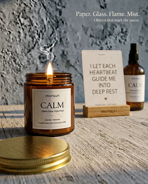 Paper, glass, flame, mist - the objects that mark the pause, with the lit CALM candle, an affirmation card in its stand and the hydrosol mist on weathered wood