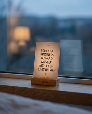 An affirmation card reading I CHOOSE KINDNESS TOWARD MYSELF WITH EACH QUIET BREATH standing on a window ledge at dusk, with the MINDFUL PAUSE deck behind it