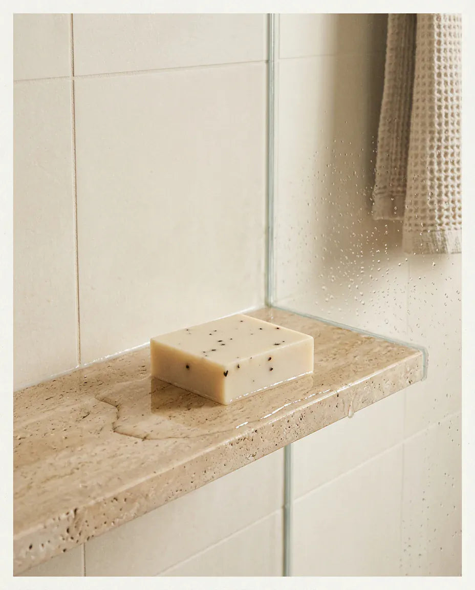 The GROUNDED cedarwood and tea tree soap bar, speckled with botanicals, resting unwrapped on a wet travertine shower ledge beside a folded waffle towel.