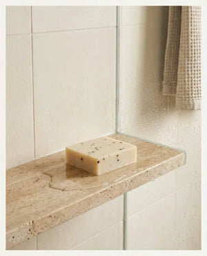 The GROUNDED cedarwood and tea tree soap bar, speckled with botanicals, resting unwrapped on a wet travertine shower ledge beside a folded waffle towel.