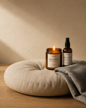 The lit BALANCE palo santo candle and the GROUNDED hydrosol mist resting on a linen meditation cushion with a grey wool blanket folded behind, in low afternoon light.