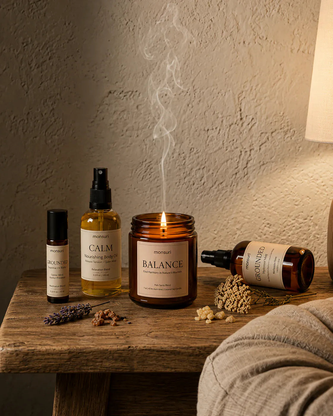 An evening bench scene lit by the burning palo santo candle — the GROUNDED meditation roller, the CALM nourishing body oil, the candle itself and the GROUNDED hydrosol laid on its side, with dried lavender and frankincense resin scattered on the wood.