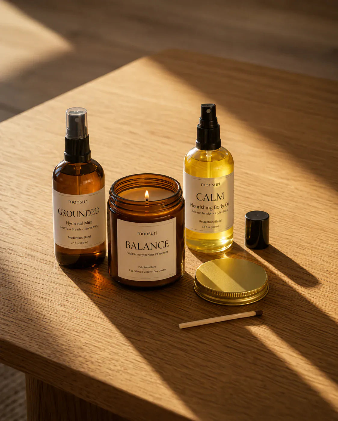 The BALANCE palo santo candle burning on an oak table between the GROUNDED hydrosol mist in amber glass and the CALM body oil in clear glass, with the candle's gold lid and a spent match set down beside them.
