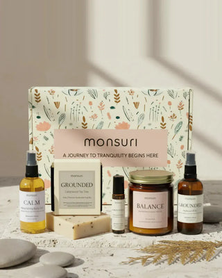 The Monsuri botanical kraft gift box with all five mindfulness pieces in front of it on pale stone — the 3.3 fl oz CALM nourishing body oil in clear glass, the 4.5 oz GROUNDED cedarwood and tea tree soap bar, the 0.33 fl oz GROUNDED meditation roller, the 7 oz BALANCE palo santo candle with its gold lid, and the 3.3 fl oz GROUNDED hydrosol mist in amber glass.