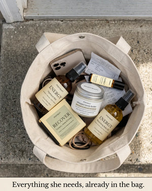 Everything she needs, already in the bag: all five pieces of the energizing gift set dropped into a canvas tote on a doorstep alongside a phone, keys and a grocery receipt — the hydrosol mist, the RECOVER soap box, the whipped body butter, the energy roller and the citrus vitality body oil.