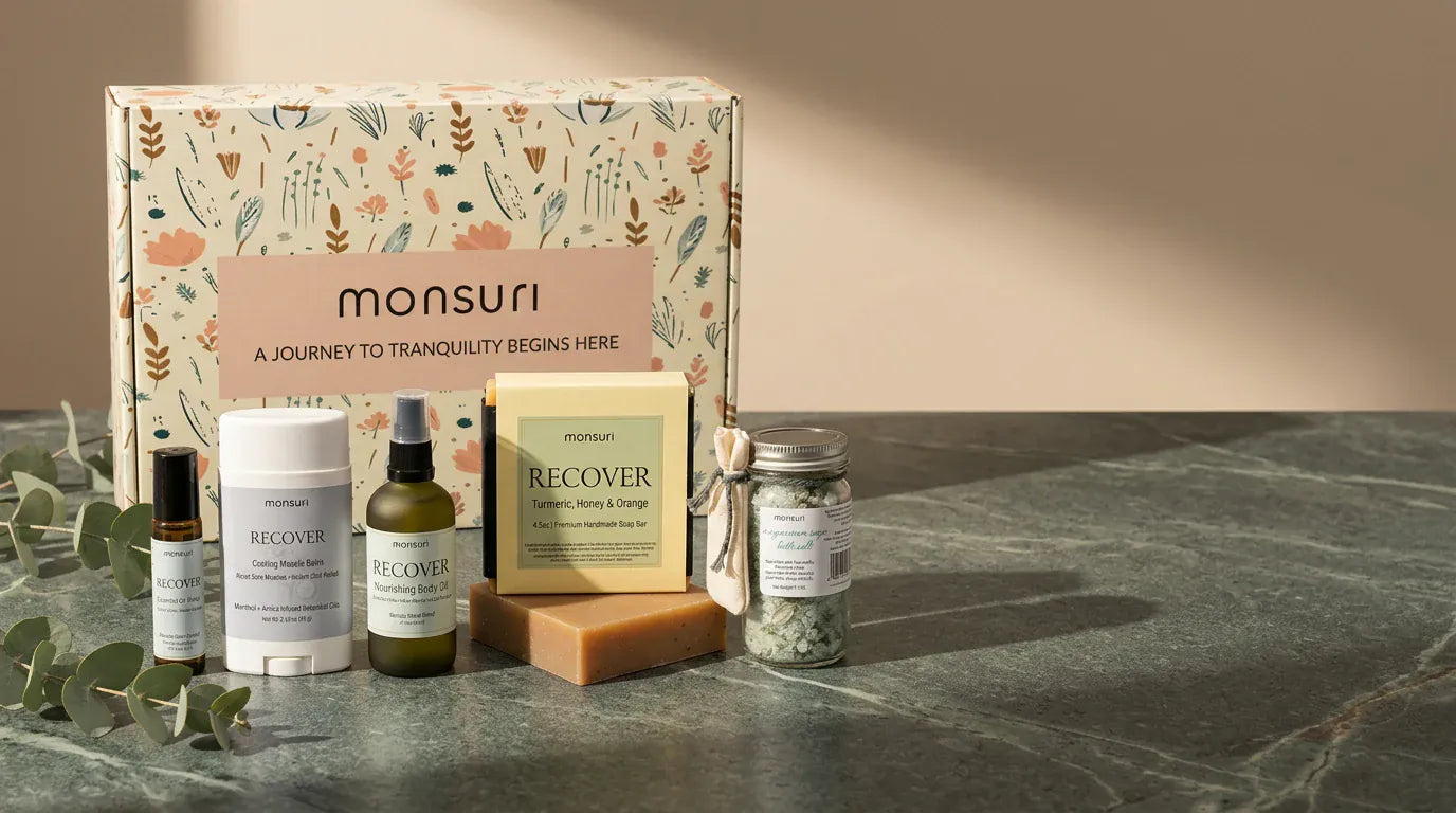 The botanical kraft gift box behind the five recovery pieces on a green marble counter in warm afternoon light — the essential-oil roller, the cooling muscle balm stick, the nourishing body oil, the boxed turmeric, honey and orange soap and the magnesium sage bath salts.