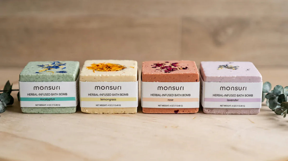 All four bath bombs lined up along a stone counter with eucalyptus at either end — eucalyptus, lemongrass, rose and lavender, each banded and labelled 4 oz (113.40 g).