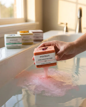 A hand lowering the rose bath bomb into a filled tub, its colour already trailing through the water, with the other three cubes lined up on the tub edge behind.