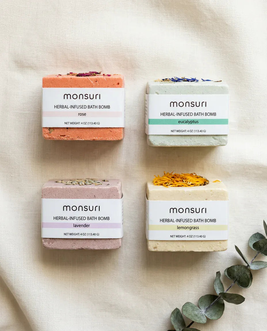 Overhead flat lay of the four bath bombs on cream linen with a eucalyptus sprig — rose, eucalyptus, lavender and lemongrass, each a 4 oz (113.40 g) cube in its paper band, with dried petals and buds pressed into the top of each.